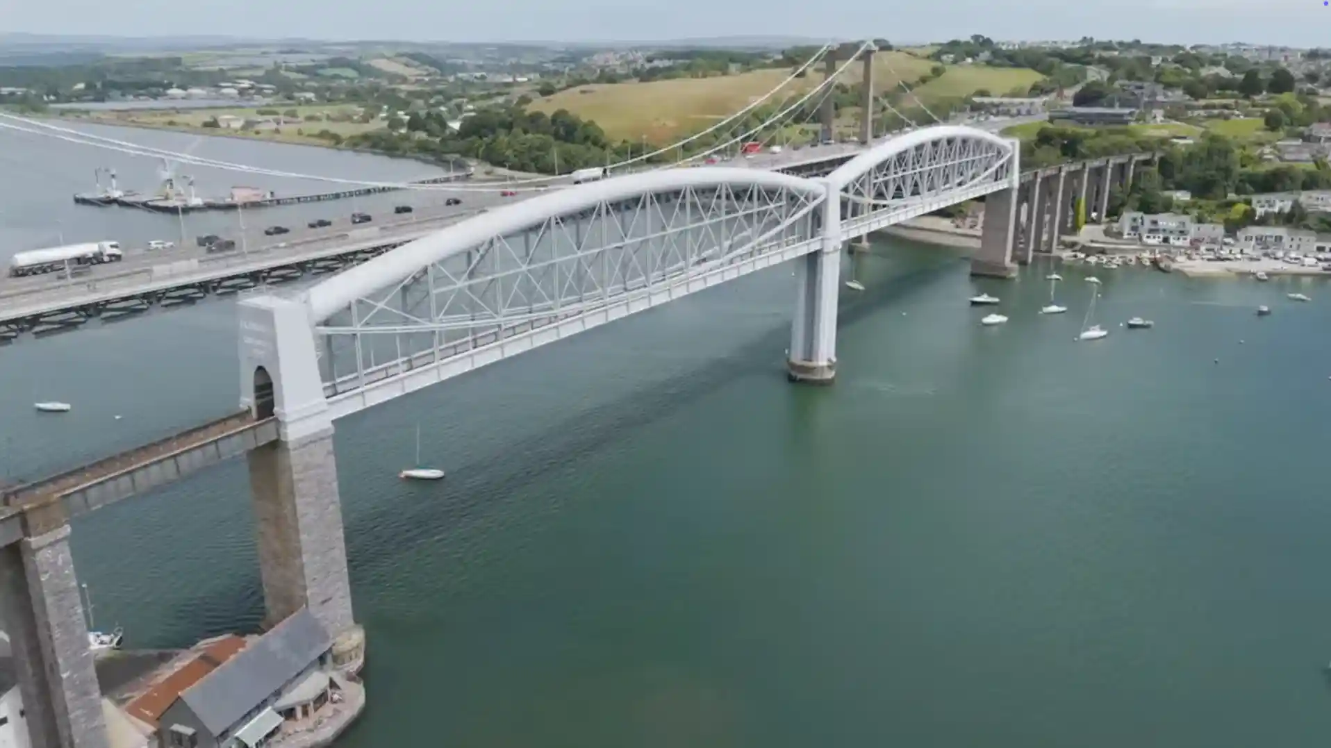 Side view of the Royal Albert Bridge and Tamar Bridge crossing the River Tamar, with boats moored below and green hills in the background.