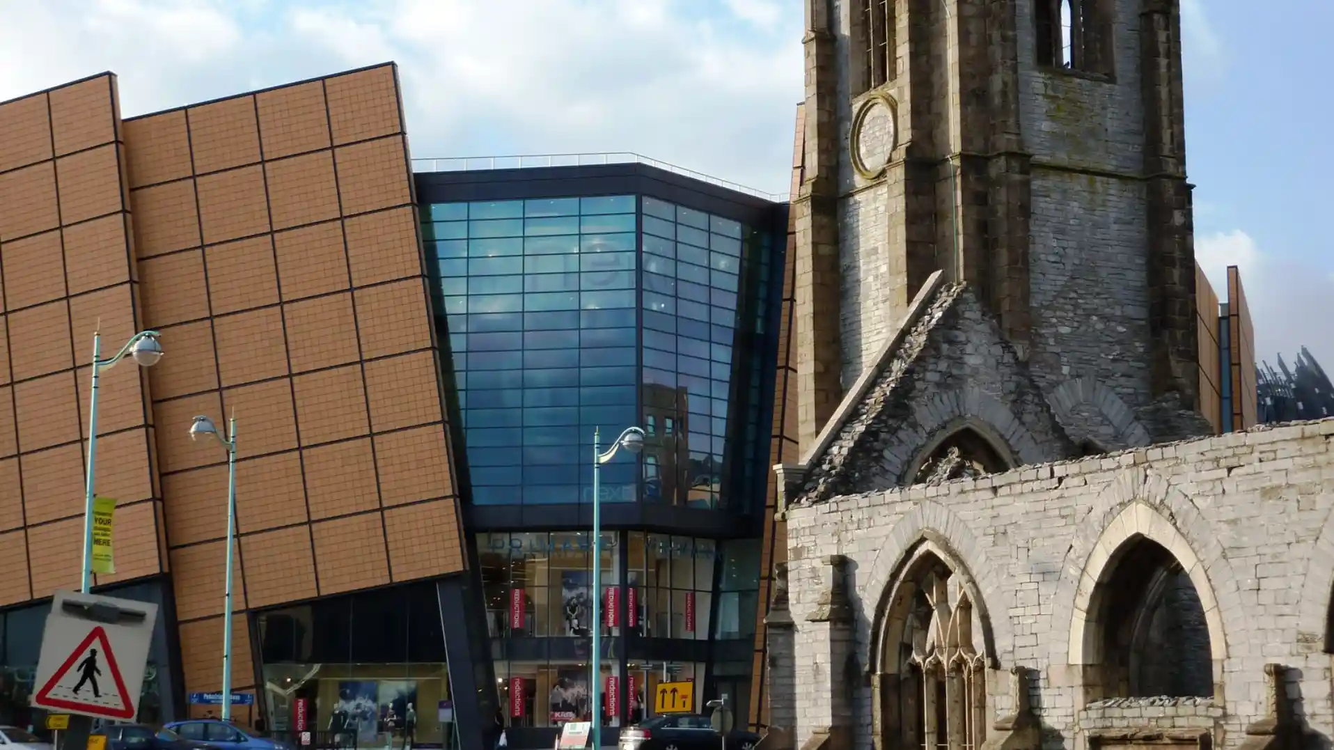 The modern glass and panelled exterior of Drake Circus shopping centre beside the stone ruins of Charles Church in Plymouth city centre.